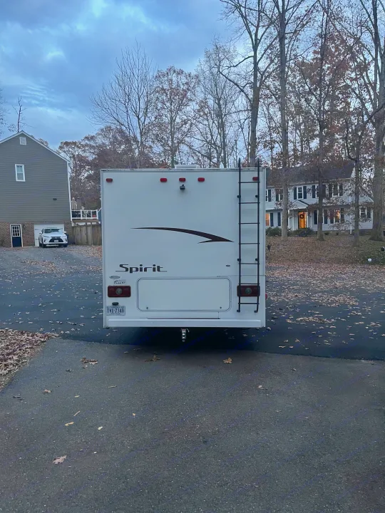 Rear view of a white Spirit RV rental, parked on an autumn driveway with fallen leaves. Houses and trees are in the background.. Winnebago Winnebago 2014