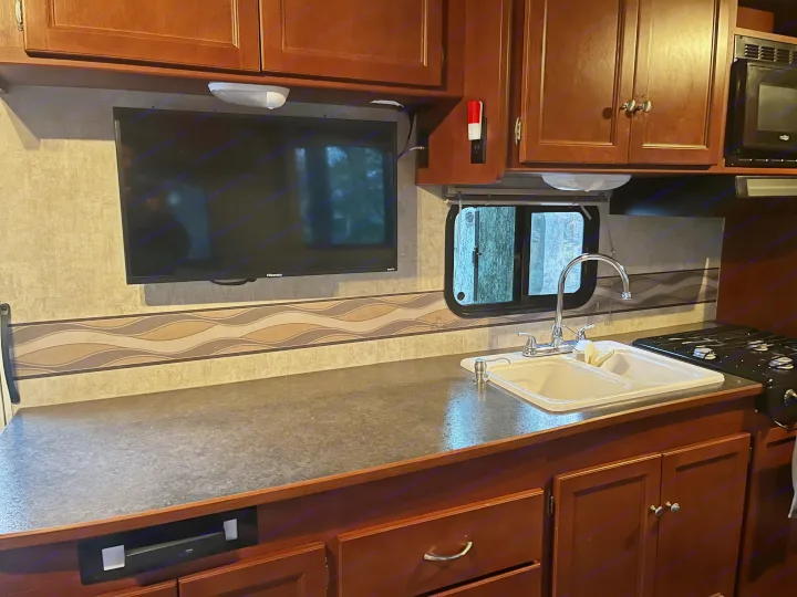 Interior of an RV rental, highlighting the compact kitchen with TV, sink, stove, microwave, & wood cabinets.. Winnebago Winnebago 2014