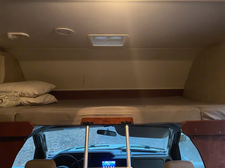 Cozy RV rental interior: cab-over bed with pillows, seen from above the front cockpit and dashboard.. Winnebago Winnebago 2014