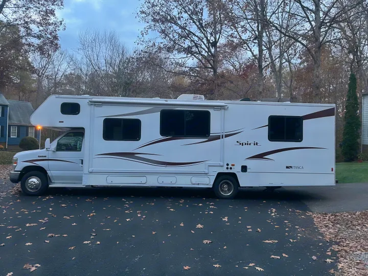 This white Class C RV rental, parked on an autumn-leaf-covered driveway, is ready for a scenic journey. "Spirit" visible on side.. Winnebago Winnebago 2014