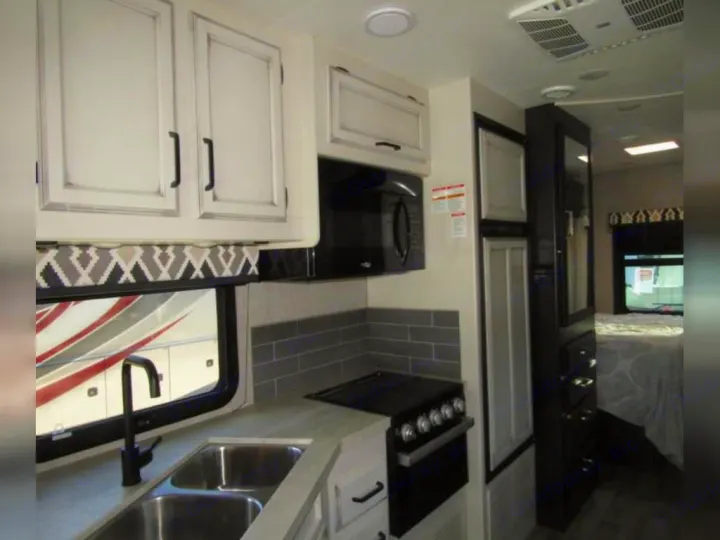 Inside an RV rental: modern kitchen with sink, stovetop, microwave, and fridge. Bedroom visible in the background.. Jayco Redhawk 2022