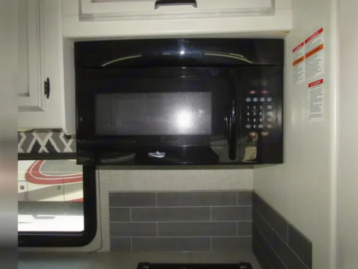 RV rental kitchen showing black microwave, gray tile backsplash, and white cabinets. A glimpse of another RV is outside the window.. Jayco Redhawk 2022
