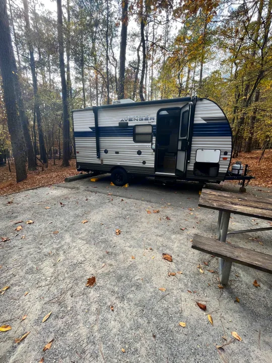 An RV rental is set up at a peaceful forest campsite with a gravel pad and picnic table, perfect for nature getaways.. Avenger LT 2022