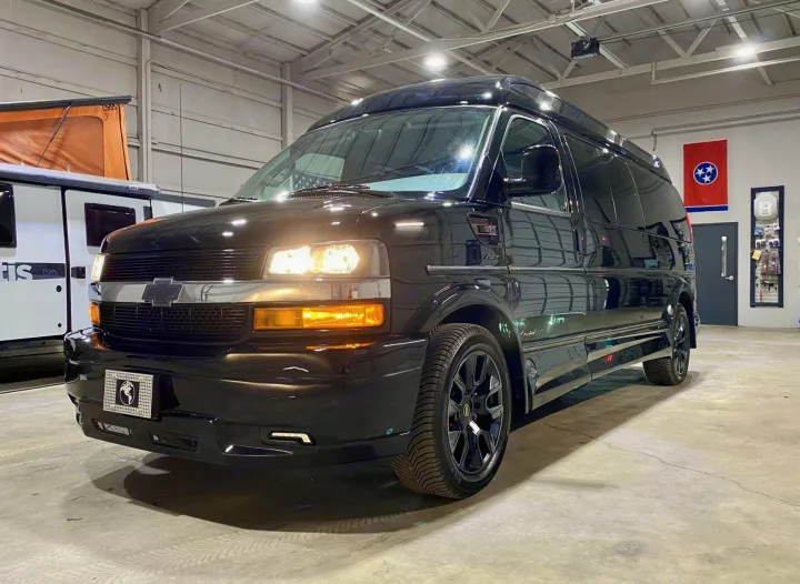 Sleek black Chevrolet conversion van, ideal for RV rental, illuminated in a clean garage with an RV trailer nearby.. Passenger Van 9 Passenger Van 2024