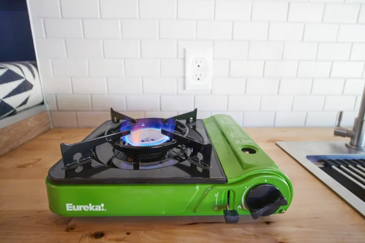 Green portable gas stove with a blue flame lit on a wooden counter in an RV rental kitchen setup.. Ford Transit 2021