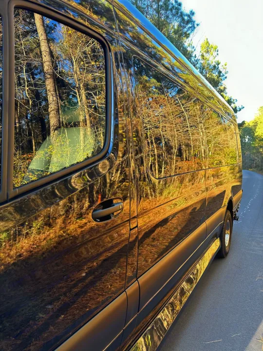 Sleek dark RV rental van reflecting a sunlit forest and blue sky, parked on a quiet road.. Ultimate Toys Ultimate Coach 2024