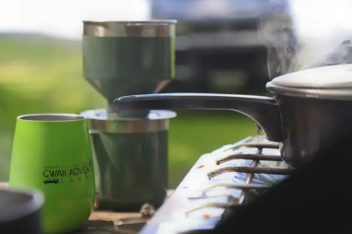Morning coffee and breakfast outdoors during an RV rental trip. Steam rises from a pot on a camp stove, next to a branded Gwaii Adventures mug.. MITSUBISHI Delica 1997