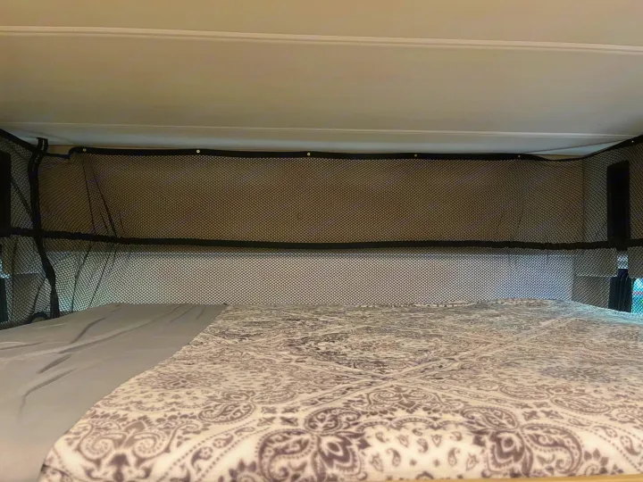 Inside an RV rental: a cozy bed with a patterned blanket and overhead mesh safety net in a sleeping bunk.. Thor Challenger 2021