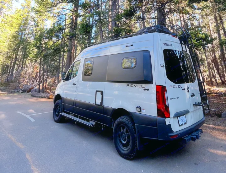 White Winnebago Revel 4x4 RV rental parked on asphalt, surrounded by tall pine trees, ready for adventure.. Winnebago Revel 2023