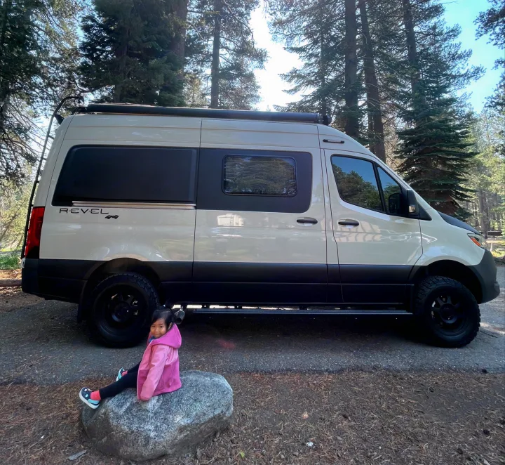 A child sits by a cream-colored Revel 4x4 camper van in a sunny forest, perfect for an RV rental adventure.. Winnebago Revel 2023