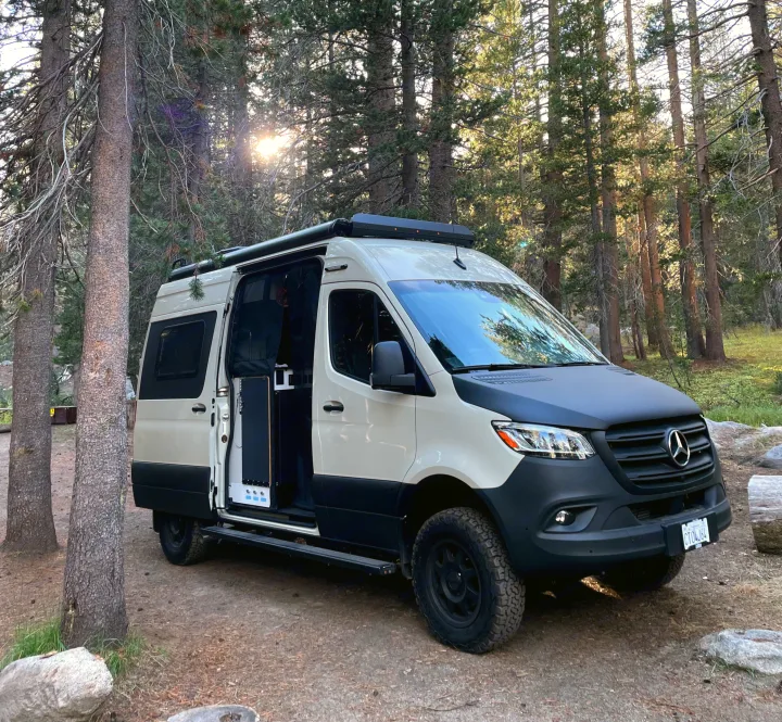 A two-tone Mercedes Sprinter camper van, likely an RV rental, is parked amidst tall trees in a sunny forest.. Winnebago Revel 2023