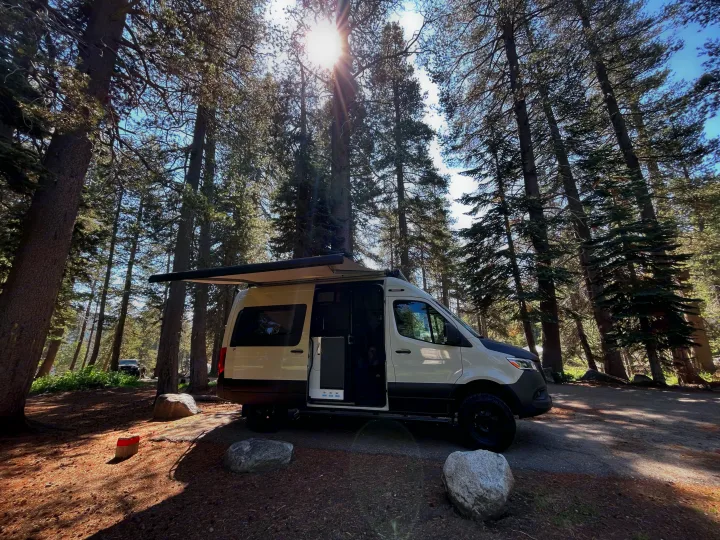 A white camper van rv rental with an extended awning is parked at a sunlit forest campsite, ready for outdoor living.. Winnebago Revel 2023