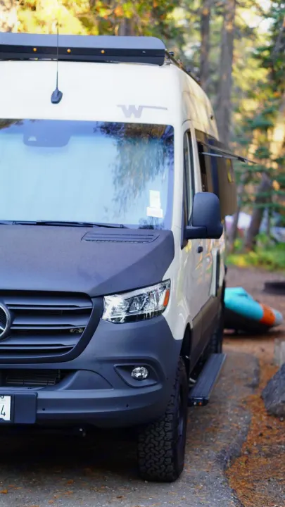A light-colored Mercedes Sprinter RV rental, modified for adventure, parked at a wooded campsite with an inflatable boat nearby.. Winnebago Revel 2023