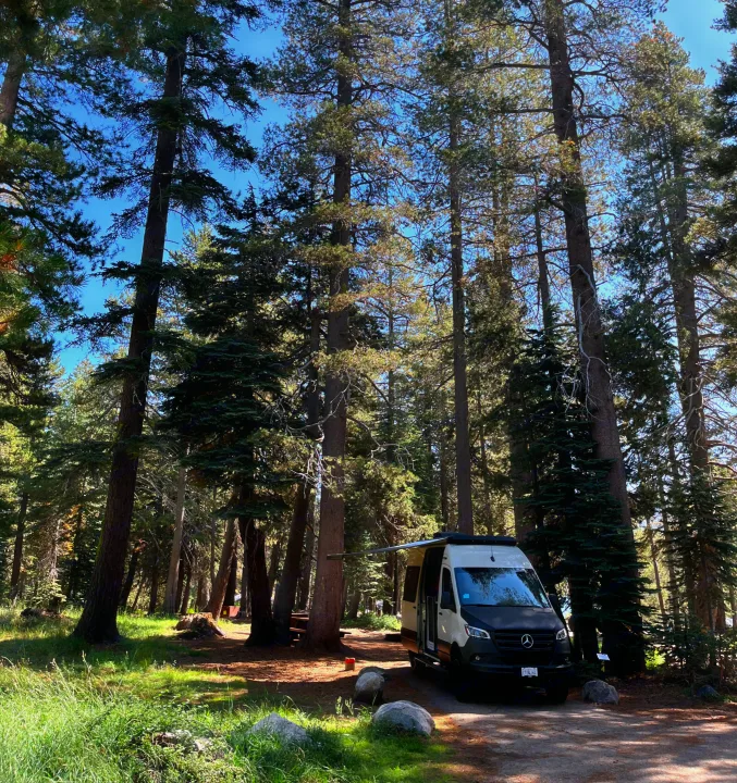 RV rental camper van parked in a sunny forest campground. Tall trees surround the peaceful campsite.. Winnebago Revel 2023