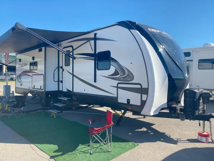 This spacious white and black RV, with its awning extended, is set up at a sunny campsite. Ideal for your next rv rental trip.. Grand Design Reflection 2022