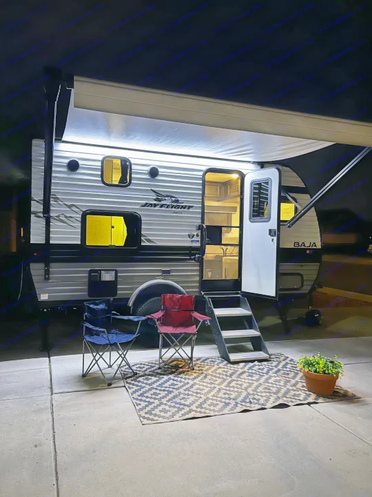 A travel trailer RV rental, illuminated at night with a lighted awning, outdoor chairs, and patterned mat set up.. Jayco Jay Flight SLX Baja Edition 2023