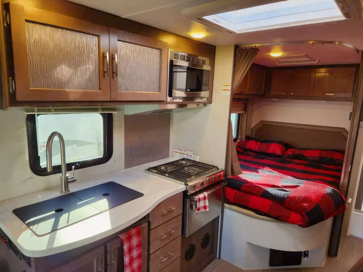 RV rental interior: A functional kitchen area with stove, microwave, and sink, alongside a cozy bedroom with plaid bedding.. Adventurer 24DS 2020