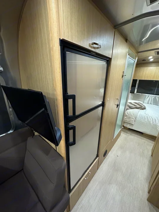 Modern RV rental interior with a Dometic fridge, TV, and sofa, leading to a compact bedroom. Light wood accents and stainless steel details are visible.. Airstream Flying Cloud 2017