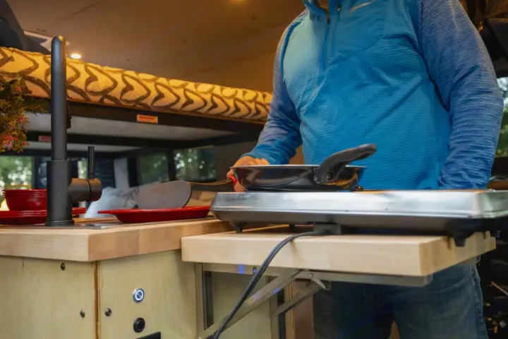 Enjoy cooking in your RV rental! A person uses an electric griddle in a camper van kitchen with a sink and bed.. Ram ProMaster 2021