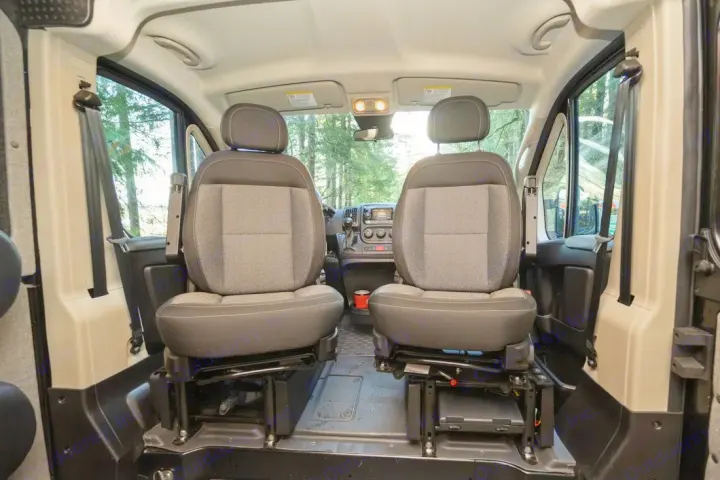 Inside an RV rental camper van, two gray and black swivel front seats face the dashboard, with a forest view ahead.. Ram ProMaster 2021