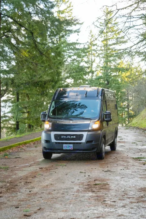 Black Ram ProMaster RV rental van on a damp forest road among tall trees, headlights on. Ideal for nature trips.. Ram ProMaster 2021