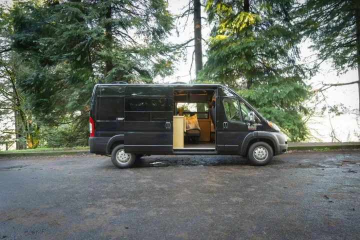 Black Ram ProMaster camper van RV rental parked among tall pine trees with side door open, showing the converted interior.. Ram ProMaster 2021