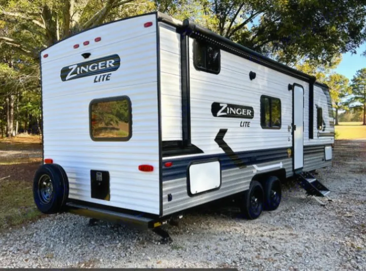 A modern white and grey Zinger Lite travel trailer, perfect for an rv rental, parked on gravel under trees.. Crossroads RV Zinger Lite 2022
