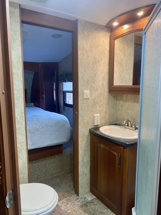 This RV rental interior features a compact bathroom with a sink, mirror, and shower, leading to a carpeted bedroom. A toilet is partly visible.. Keystone Cougar 2012