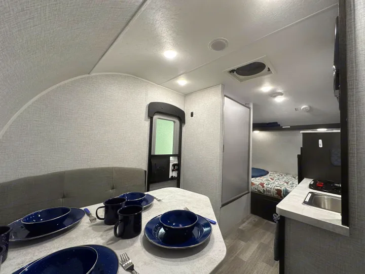 Step inside this RV rental's inviting interior: curved walls, a dining area set with blue dishes, and a compact kitchen next to cozy bunk beds.. Braxton Creek Bushwhacker Plus 2023