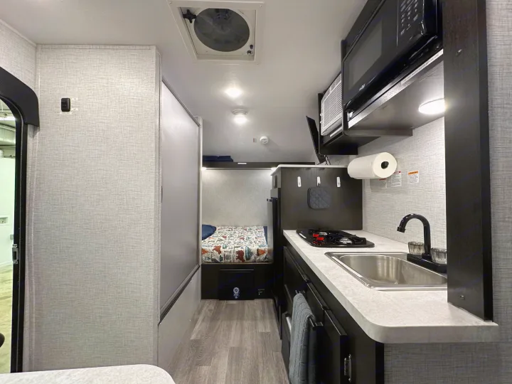 Cozy interior of an RV rental, featuring a compact kitchen with a sink and stove, and a comfortable bed area.. Braxton Creek Bushwhacker Plus 2023