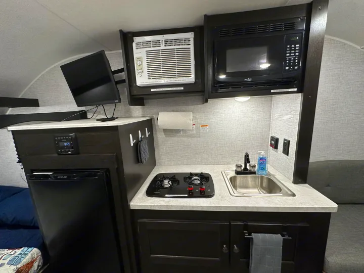 Inside this RV rental, a compact kitchen features a sink, two-burner stove, microwave, AC, TV, and small fridge.. Braxton Creek Bushwhacker Plus 2023