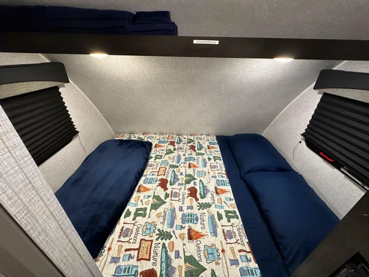 RV rental's cozy sleeping area: a bed with a camping-themed quilt, navy bedding, and overhead storage with a 15 lb limit.. Braxton Creek Bushwhacker Plus 2023