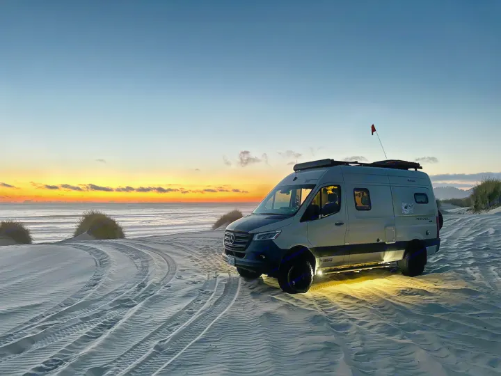 Stunning beach RV rental adventure! A camper van sits on sandy dunes at a vibrant sunset by the ocean, lights illuminating the sand.. Winnebago Revel 2020