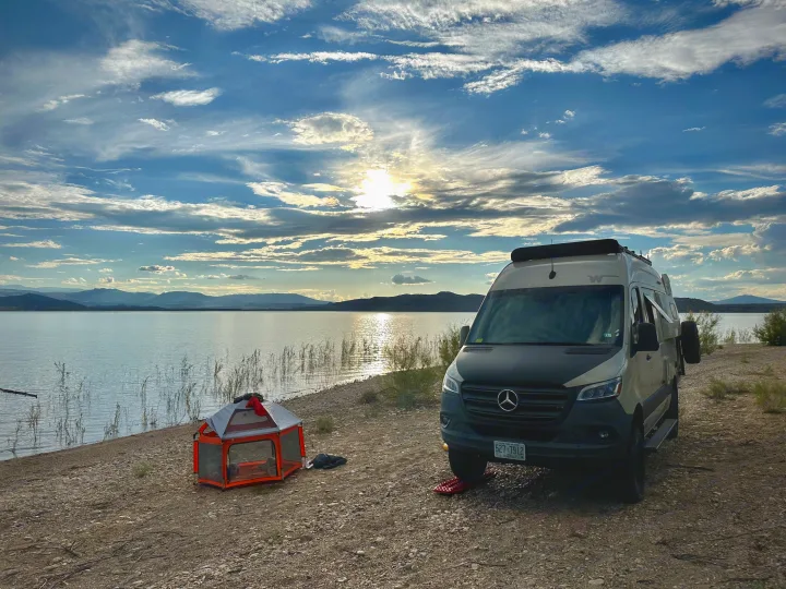 RV rental camper van parked by a scenic lake, mountains in distance, and an orange playpen on shore at sunset.. Winnebago Revel 2020
