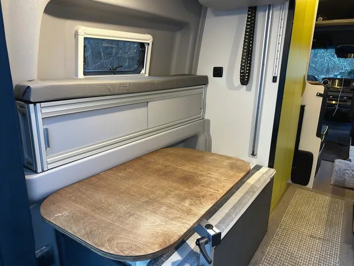 RV rental interior: A compact camper van featuring a bed, window, wooden table, and integrated storage for versatile mobile living.. Winnebago Revel 2020