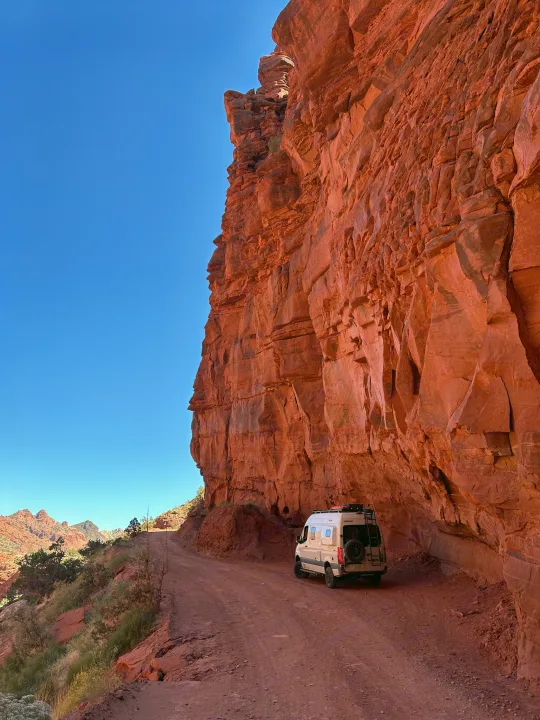 RV rental on a rugged dirt road, dwarfed by a massive red rock cliff under a clear blue sky, perfect for desert adventure.. Winnebago Revel 2020