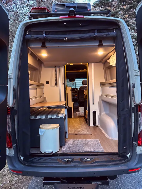 Looking into a cozy camper van rv rental with open rear doors, featuring a bed, table, and ambient lighting.. Winnebago Revel 2020
