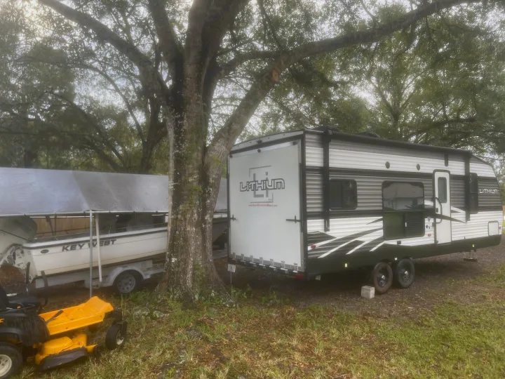 A gray RV rental and a boat are parked next to a large tree, with a yellow riding mower nearby on a cloudy day.. Heartland Lithium 2021
