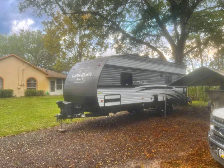 A grey and white Lithium RV rental (travel trailer) is parked on a gravel drive next to a house and large trees.. Heartland Lithium 2021