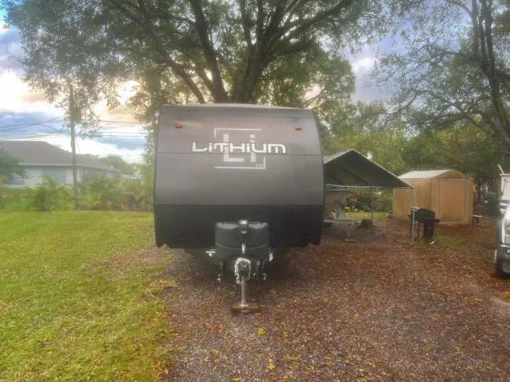 Dark gray LiTHIUM RV rental on gravel, surrounded by trees and grass, with a house and shed in the background.. Heartland Lithium 2021