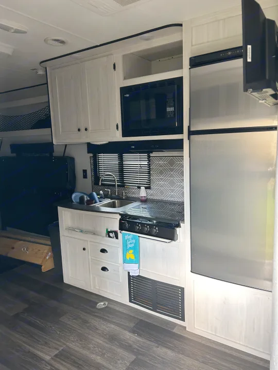 Compact rv rental kitchen: sink, stove, microwave, and fridge, with light wood cabinets and dark flooring.. Heartland Lithium 2021