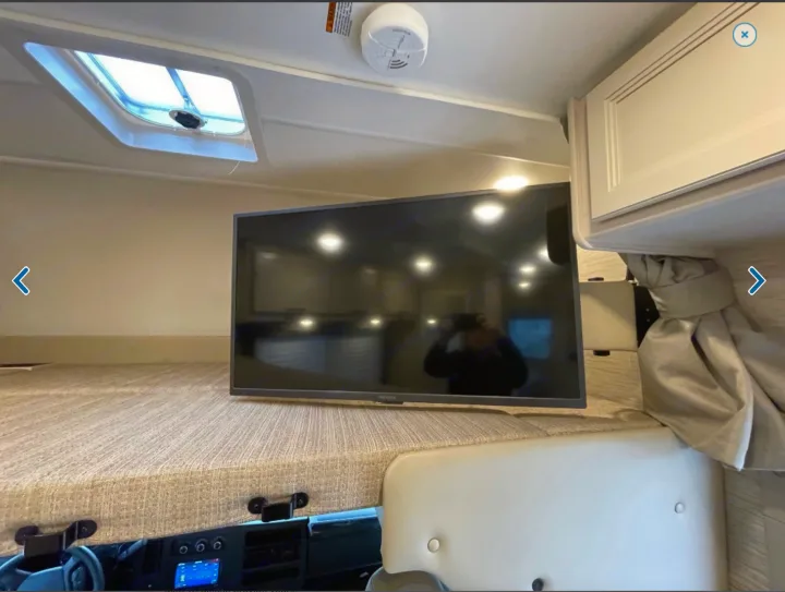 This RV rental features a cozy overhead bunk with a mounted TV, skylight, and dashboard view, offering a functional sleeping area.. Thor Freedom Elite 2023