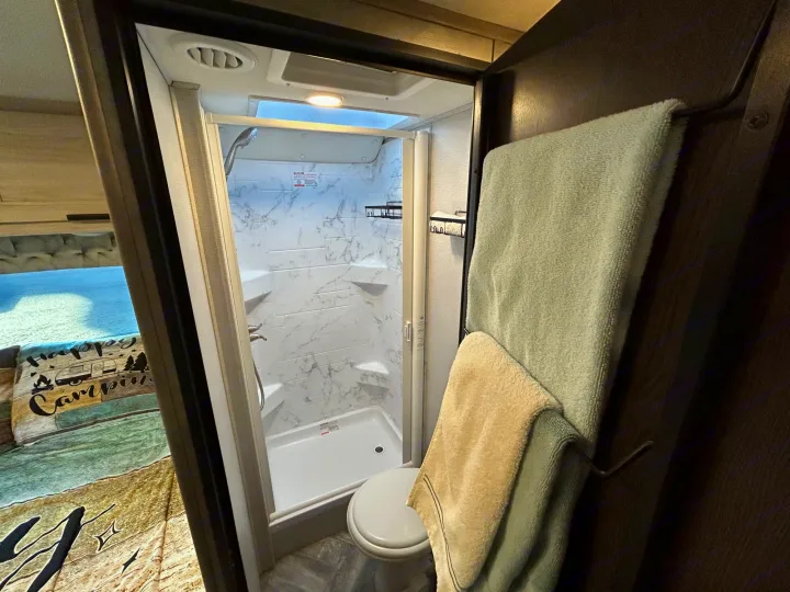 This rv rental features a compact bathroom with shower, toilet, and an adjacent bedroom area, complete with towels.. Forest River Sunseeker LE 2022