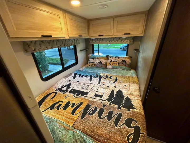 Cozy rv rental bedroom featuring a "Happy Camping" bed, overhead cabinets, and large windows. Perfect for your next adventure!. Forest River Sunseeker LE 2022