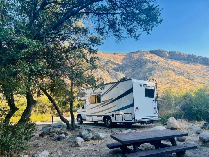 A white Sunseeker RV rental is parked at a scenic campsite with a picnic table, surrounded by trees and mountains in the background.. Forest River Sunseeker LE 2022