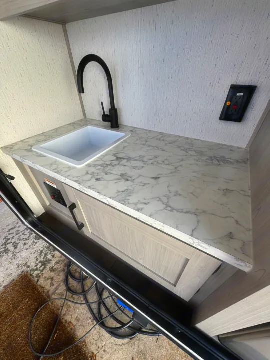 RV rental outdoor kitchen featuring a black faucet, white sink, and marble countertop. Power cords visible outside.. Dutchmen Other 2023