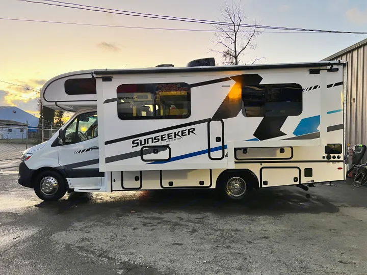 Side view of a modern white Sunseeker **RV rental** with blue and black graphics, parked on pavement at sunset.. Forest River Sunseeker 2024