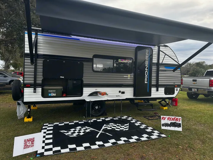 An RV rental travel trailer with an extended awning and "RV RENTALS" sign is set up on a checkered mat outdoors.. Heartland RVs Pioneer 2024
