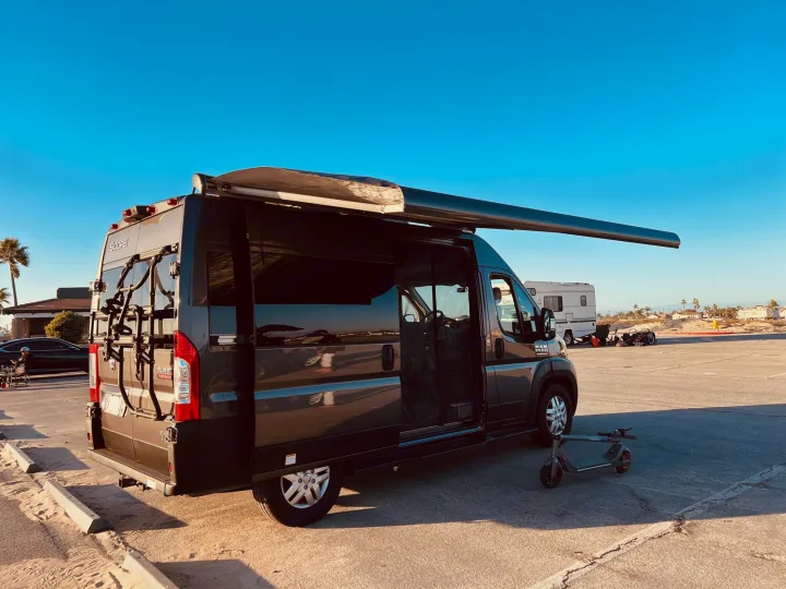Grey camper van with extended awning, bike rack, and scooter. Perfect for an RV rental beach adventure under a clear sky.. Thor Scope 2023
