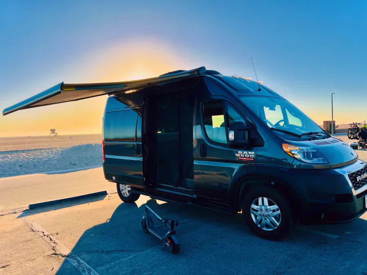 This Ram ProMaster camper van, perfect for rv rental adventures, is parked by the beach with its awning extended and door open. An e-scooter is nearby, ready for exploration.. Thor Scope 2023